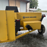 Yellow New Holland agricultural machine with a black vent panel, situated on a concrete yard near a barn and trailer in the background.