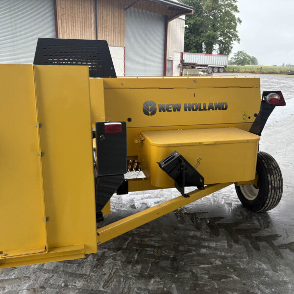 Yellow New Holland agricultural machine with a black vent panel, situated on a concrete yard near a barn and trailer in the background.