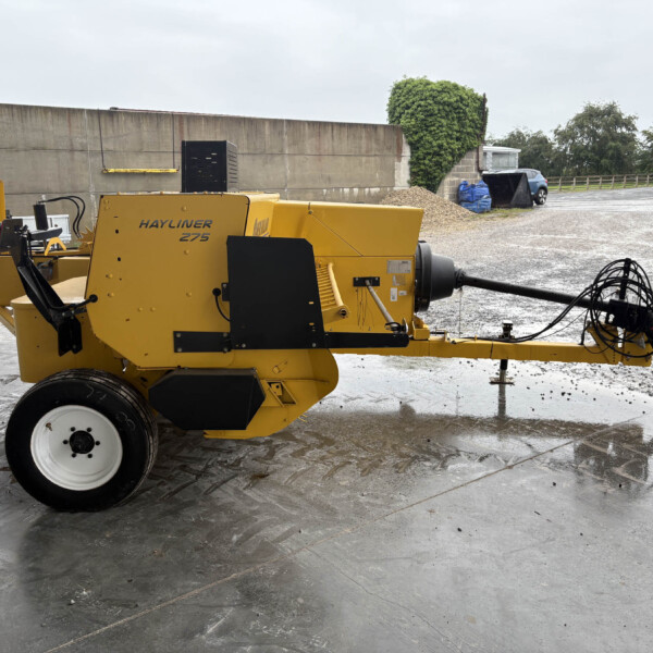Yellow Hayliner 275 baler in a wet outdoor yard with a gravel pile and a blue car in the background.