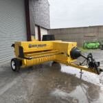 Yellow New Holland Hayliner 275 hay baler parked outside beside a corrugated metal building, with wet ground and scattered equipment nearby.