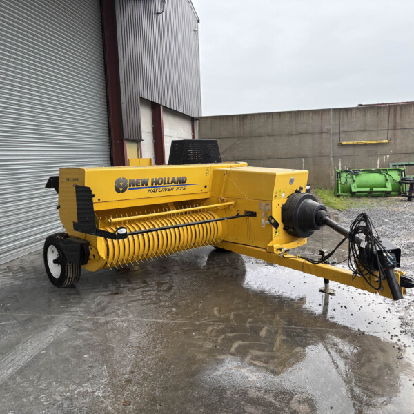 Yellow New Holland Hayliner 275 hay baler parked outside beside a corrugated metal building, with wet ground and scattered equipment nearby.