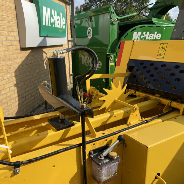 Close-up of a green McHale baler with yellow safety guards beside a brick building.
