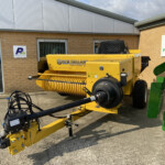 Yellow New Holland hay baler outside a brick building with a Russell Group sign visible nearby.