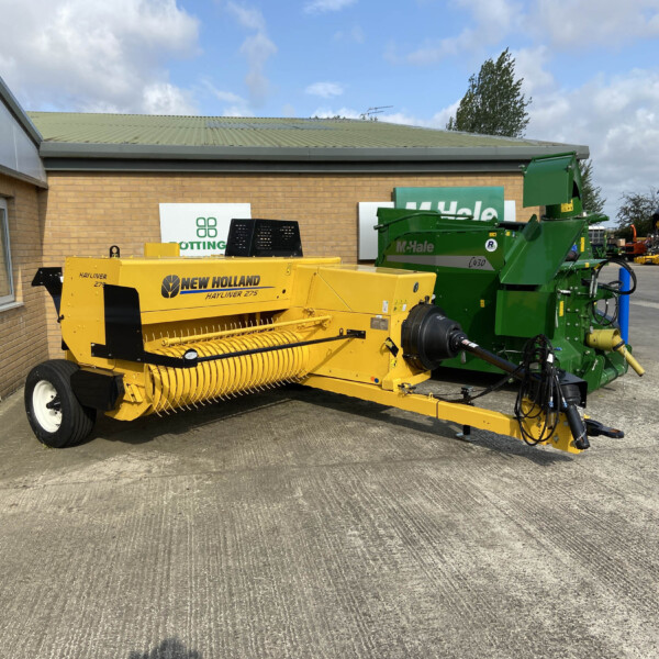 Yellow New Holland hay baler parked outside a brick building with a green McHale baler beside it, under a partly cloudy blue sky.