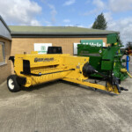 Outdoor yard with yellow New Holland Hayliner 275 hay baler next to a brick building and green McHale baler in the background under a blue sky.