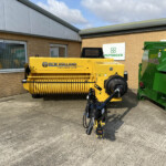 Yellow New Holland Hayliner 275 baler parked outside a brick building with company signs, including Pottinger and Russell Group logos, under a blue sky.