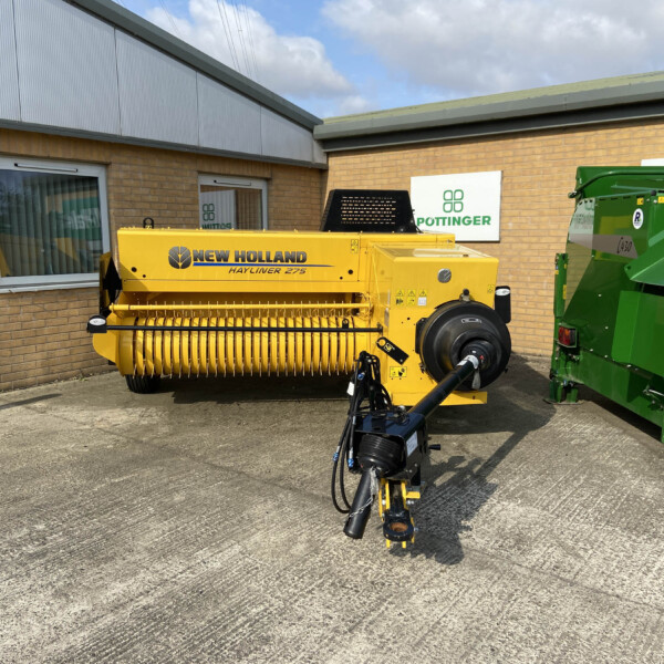 Yellow New Holland Hayliner 275 baler parked outside a brick building with company signs, including Pottinger and Russell Group logos, under a blue sky.