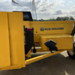 Close-up of a yellow New Holland machine on a trailer, showing branding and a wheel in an outdoor lot.