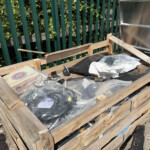 Disorganized outdoor pallet stack with plastic-wrapped debris, metal pieces, and fabric, beside a green fence and foliage in the background.