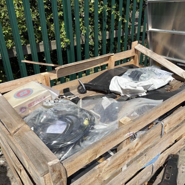Disorganized outdoor pallet stack with plastic-wrapped debris, metal pieces, and fabric, beside a green fence and foliage in the background.