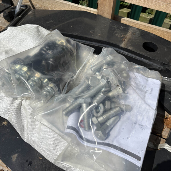 Bagged hardware parts (bolts, nuts, and spacers) placed on a dark surface next to a wooden railing and a light-colored strap or fabric edge.