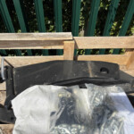 Top-down view of a weathered wooden pallet with a black plastic container and a clear bag of metal hardware, set against a green vertical fence background.