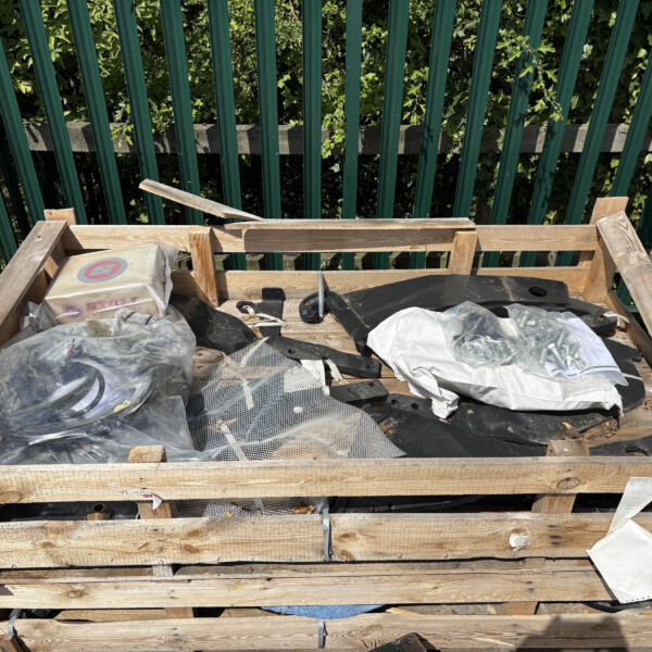 Wooden pallet with construction debris and bags on a work platform, green fence and foliage behind.