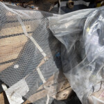 Piles of construction debris including white plastic mesh, wood planks, and a rusted pipe among a torn tarp or plastic sheeting.