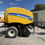 Yellow New Holland Roll-Belt 150 bale baler in an outdoor yard beside a white trailer, under a blue sky with clouds, ready for use in farming.