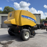 Yellow New Holland Roll-Belt 150 combine harvester on trailer in a yard with a metal building and blue sky.