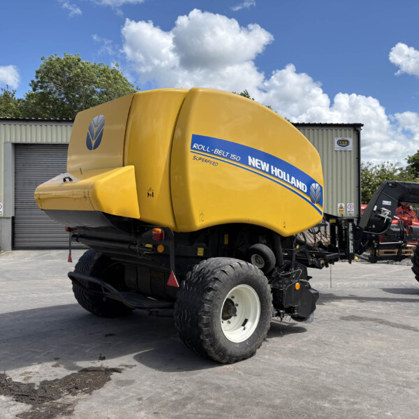 Yellow New Holland Roll-Belt 150 combine harvester on trailer in a yard with a metal building and blue sky.