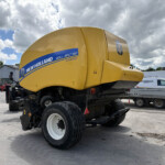 Yellow New Holland Roll-Belt 150 baler on a trailer in an outdoor yard with other farming equipment and a cloudy sky behind it.