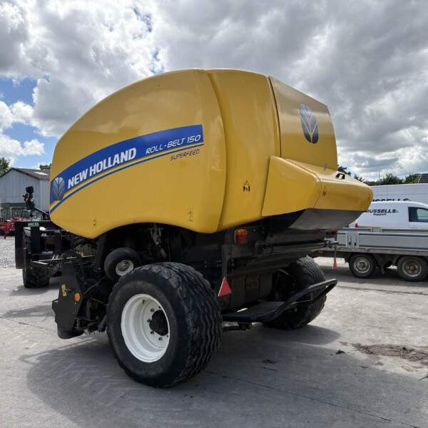 Yellow New Holland Roll-Belt 150 baler on a trailer in an outdoor yard with other farming equipment and a cloudy sky behind it.
