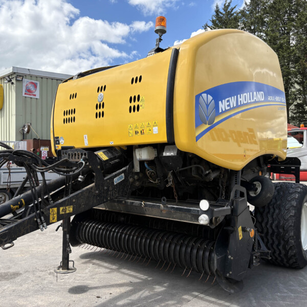Yellow New Holland agricultural machine with a header attached, parked in a lot with signs in the background reading New Holland Agriculture.