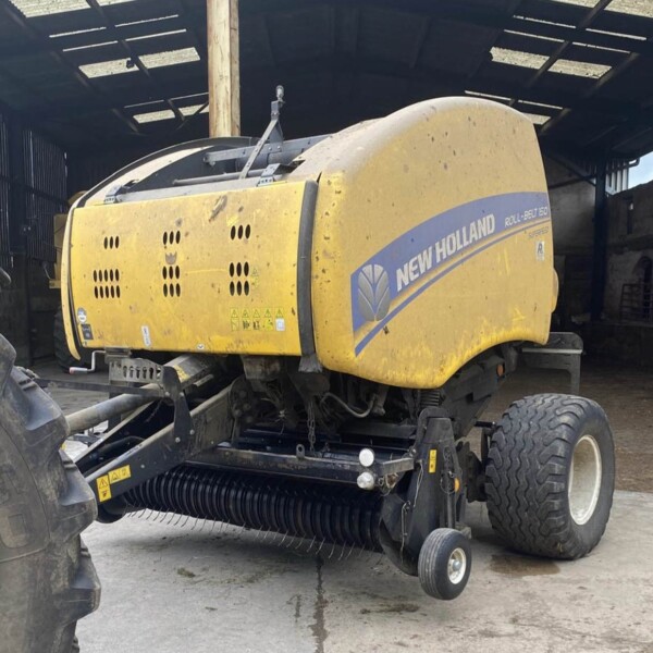Yellow New Holland round baler parked in a shed, logo visible on the side, with tractor tires nearby and hay-baling components underneath.