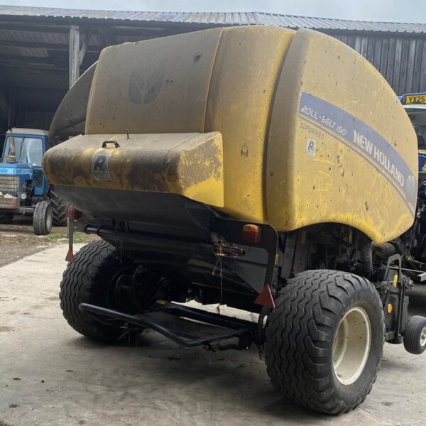 Yellow New Holland Roll-Belt 150 baler hitched to a tractor in a dusty farmyard shed, with other blue equipment nearby