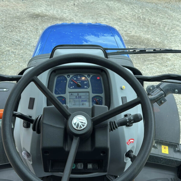 View from tractor cab showing steering wheel, dashboard, and blue hood outside on gravel ground