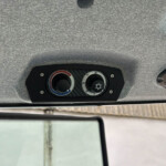Overhead vehicle ceiling with climate control panel featuring two circular dials and two dome lights on a gray fabric headliner.