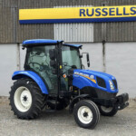 Blue New Holland tractor parked in front of a corrugated gray wall with a yellow and blue Russell's sign above it.