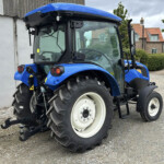 Blue agricultural tractor parked on a gravel driveway, showing large rear tires and a hitch at the back with a rural house and hedge in the background