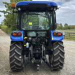 Rear view of a blue agricultural tractor on a gravel road, showing hydraulic connections and hitch at the back, with large tires and orange beacon light on the left