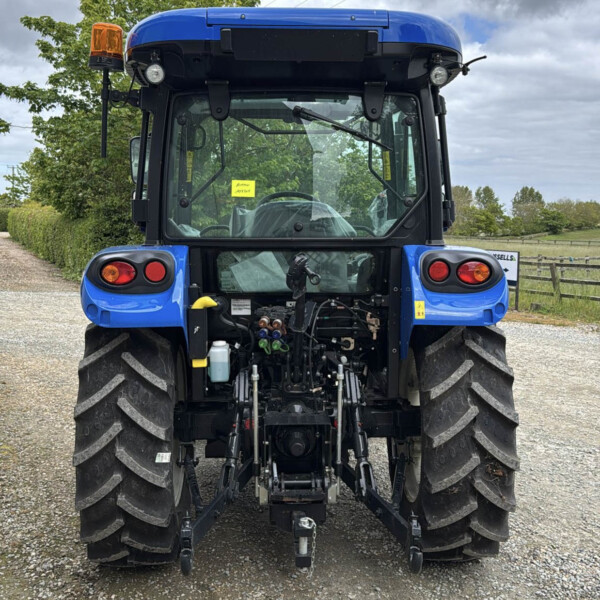Rear view of a blue agricultural tractor on a gravel road, showing hydraulic connections and hitch at the back, with large tires and orange beacon light on the left
