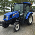 Blue New Holland tractor on a gravel lot with other tractors and farm buildings in the background.