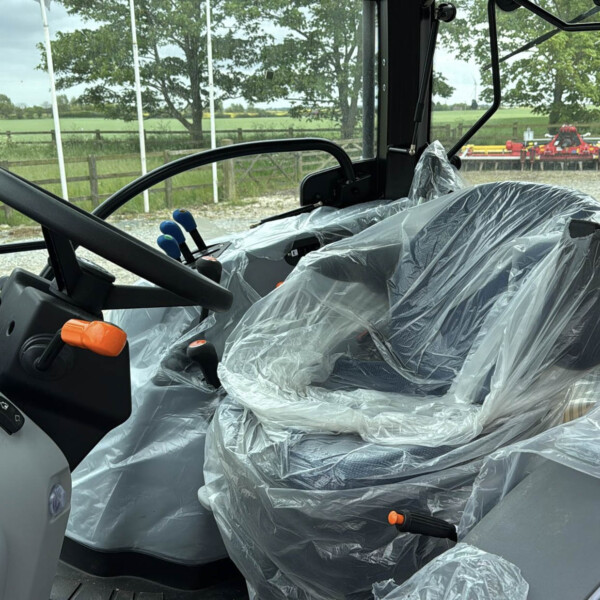 Interior of a tractor cab with plastic-covered seat, steering wheel, and control levers; rural field visible through the windows.