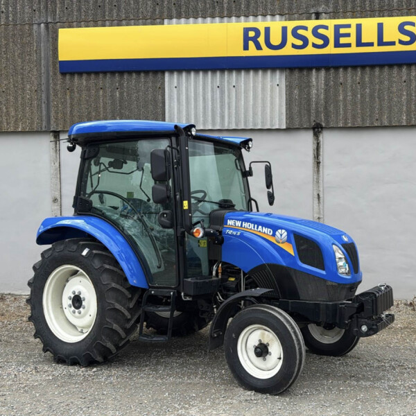 Blue New Holland tractor parked in front of a corrugated gray wall with a yellow and blue Russell's sign above it.
