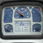 Dashboard of a vehicle showing a central RPM gauge, digital display, and warning icons on blue panels.