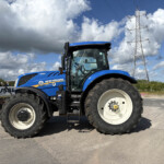 Blue New Holland tractor in a field with a tall transmission tower and power lines under a cloudy sky.