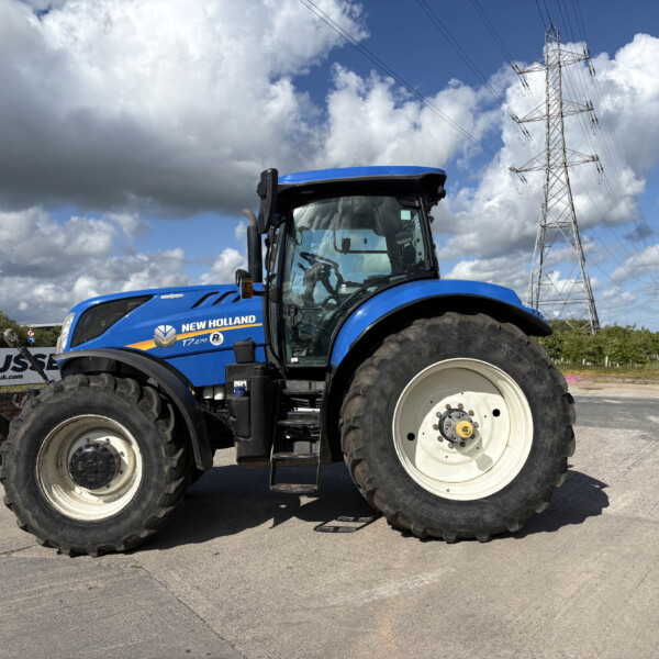 Blue New Holland tractor in a field with a tall transmission tower and power lines under a cloudy sky.