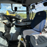 Inside view of a tractor cab with steering wheel, blue seat, and control panel; equipment visible outside the windshield on a sunny day