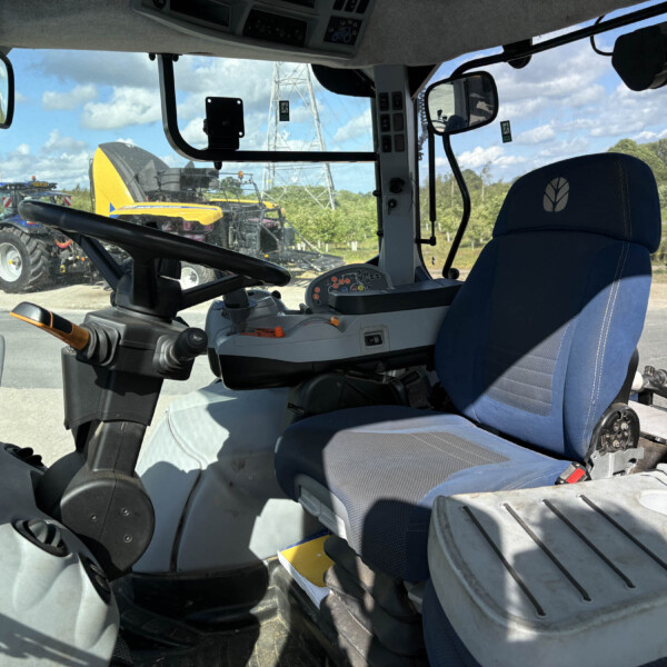 Inside view of a tractor cab with steering wheel, blue seat, and control panel; equipment visible outside the windshield on a sunny day