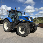 Blue New Holland tractor on a display lot with oversized tires, power lines overhead, and a Russell sign in the background