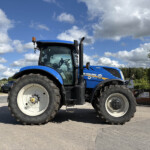Blue New Holland tractor seen from the front with oversized tires and an orange warning beacon, in a parking lot under a cloudy sky.