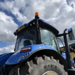 Blue tractor front view with an orange beacon on top against a cloudy sky. Large treaded tires are visible at the bottom corner.