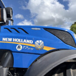 Close-up of a blue New Holland tractor cab showing the brand logo, orange side marker light, and surrounding black exhaust and panels against a blue sky.