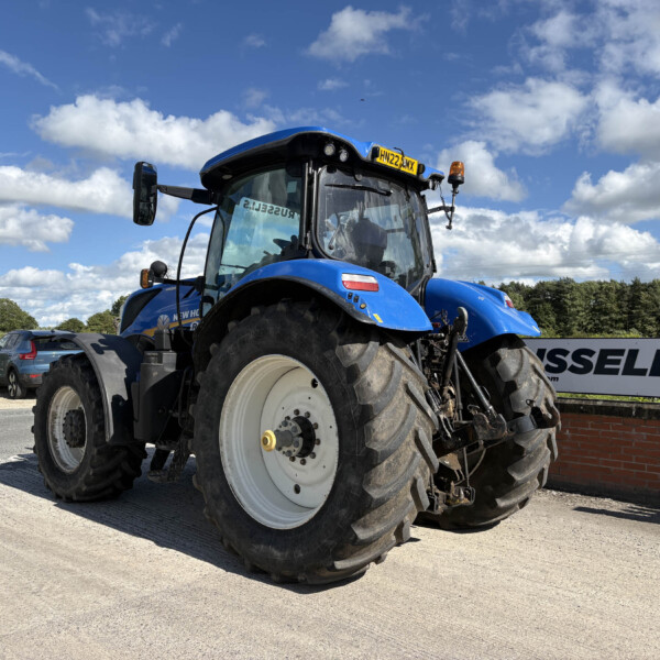 Front three-quarter view of a blue New Holland tractor with a large front tire, parked near a brick wall.