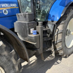 Close-up side view of a blue New Holland T7.270 tractor, showing a large rear tire, cab window, and hydraulic reservoir.