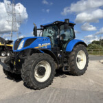 Front-right view of a blue New Holland tractor with oversized tires, parked in a lot with power lines and farm equipment in the background.