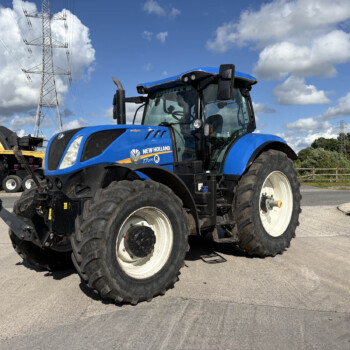 Front-right view of a blue New Holland tractor with oversized tires, parked in a lot with power lines and farm equipment in the background.