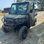 Gray Polaris Ranger off-road utility vehicle parked on a gravel lot, with stacked blocks and farm equipment visible in the background.