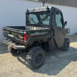 Dark green Polaris Ranger diesel utility vehicle with an empty cargo bed parked on gravel beside a white wall, outdoors.